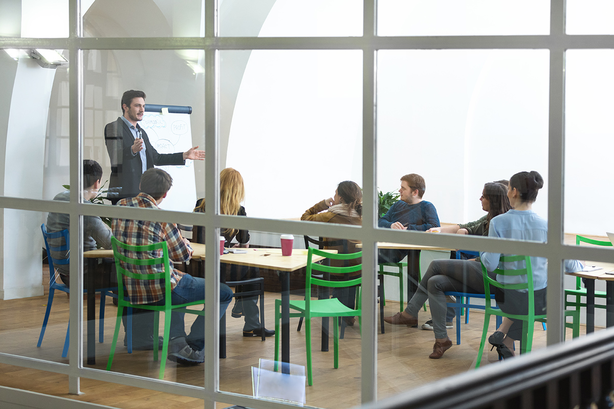 People sitting attentively in a glass-walled room, with a man talking to them in front of a flipchart